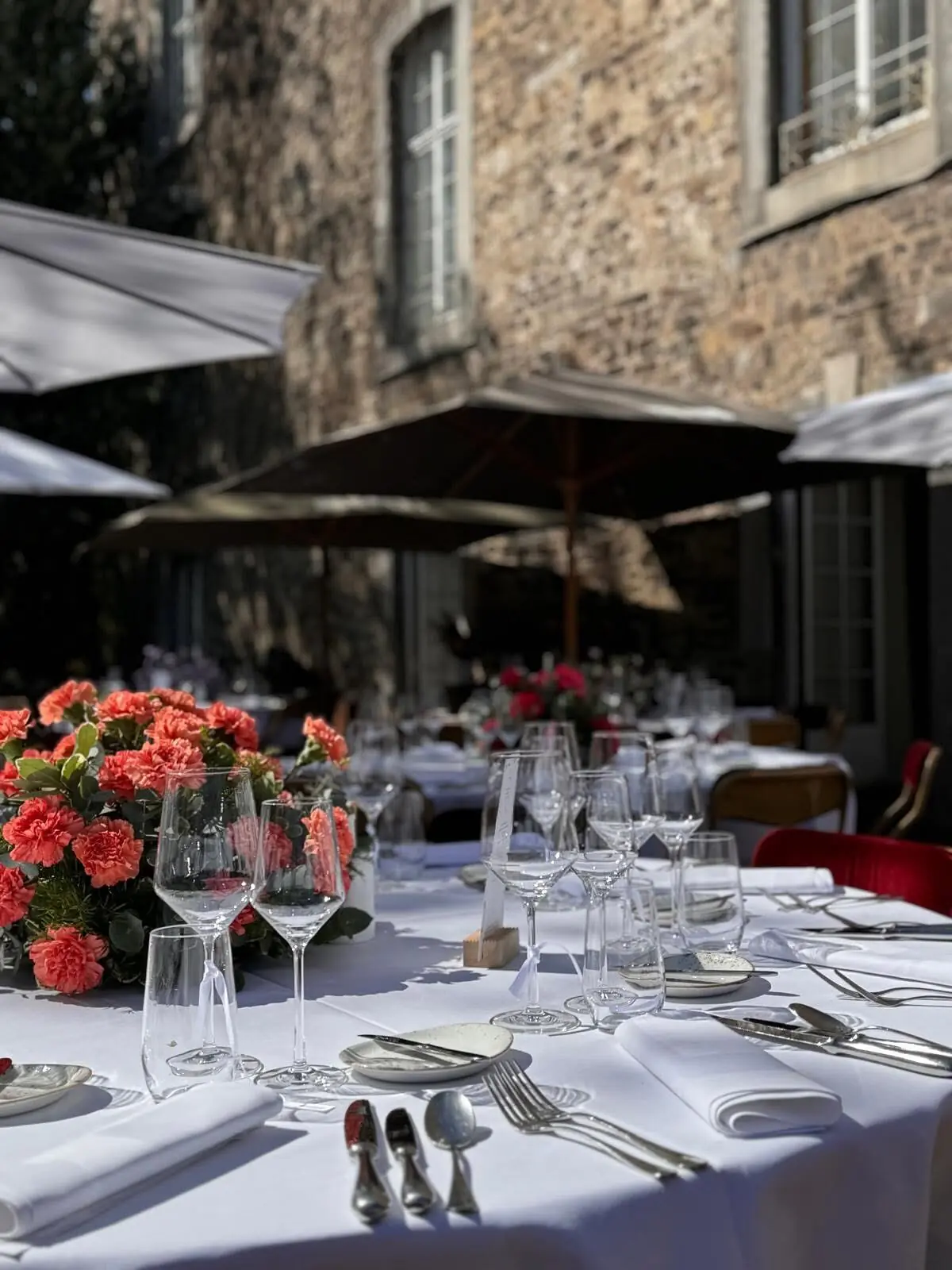 Tables dressées en terrasse, verres et fleurs, sous les arbres.