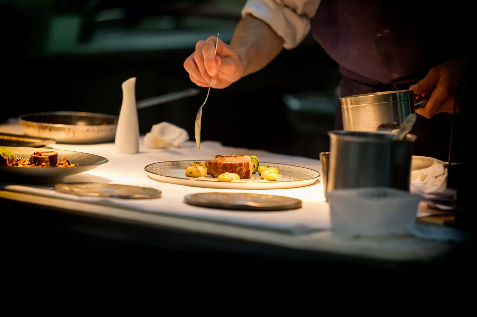 Un cuisinier nappe une assiette de sauce, sous la lampe du passe.